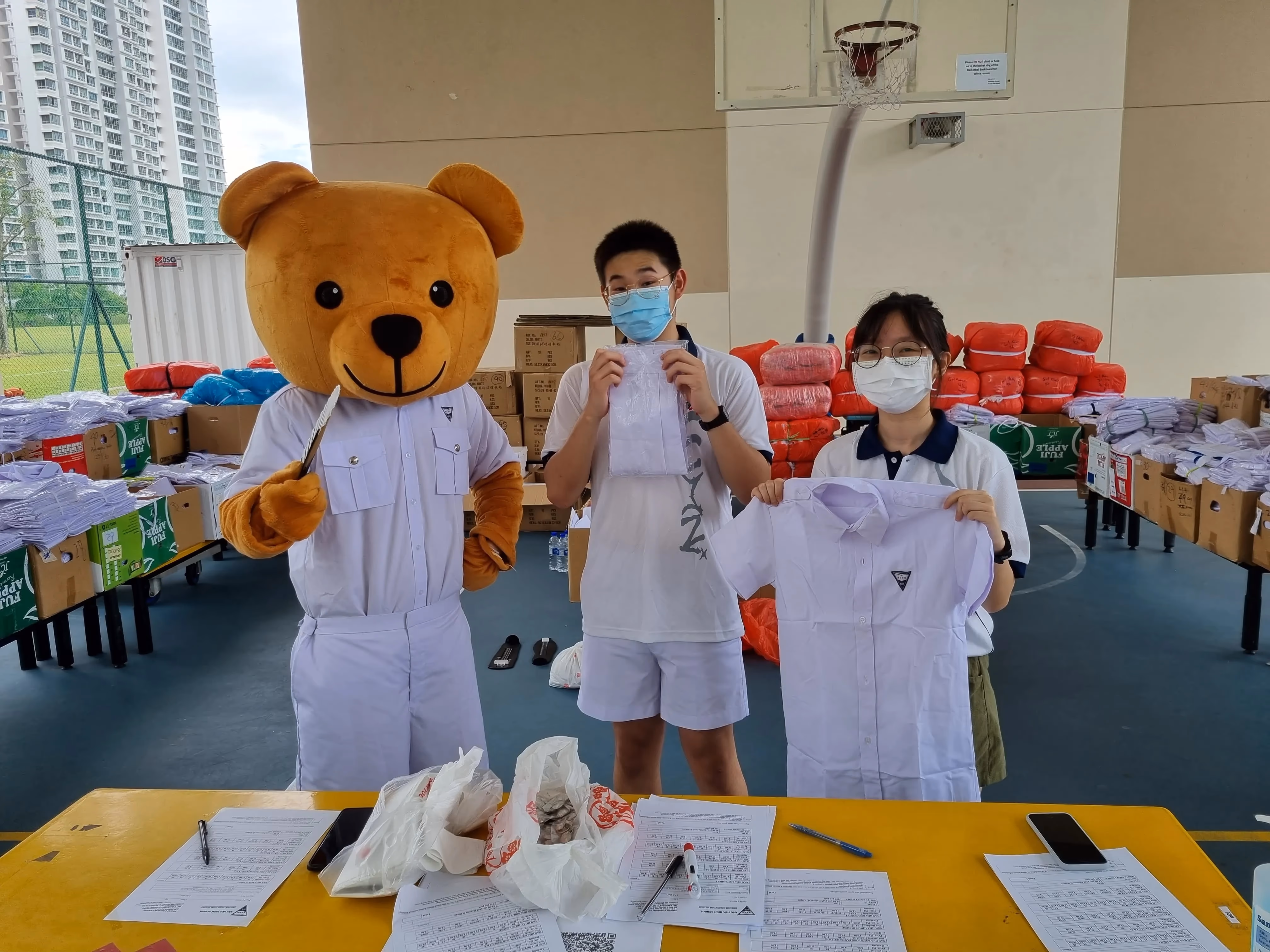 Two masked students hold up school uniforms beside a person in a brown bear mascot costume in a gym.
